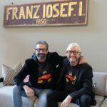 Mauro Maloberti und Franz‑Josef Baur sitzen gemeinsam auf einem Sofa unter einem Schild mit der Aufschrift 'Franz Iosef I.'.