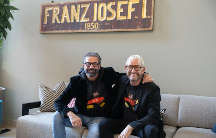 Mauro Maloberti und Franz‑Josef Baur sitzen gemeinsam auf einem Sofa unter einem Schild mit der Aufschrift 'Franz Iosef I.'.